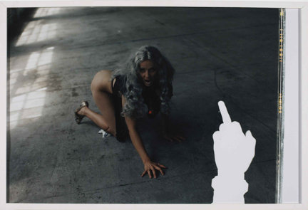framed photograph of artist in long wig crouching on the floor with a cut out hand with middle finger up